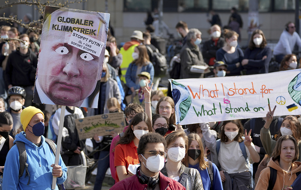 A crowd of people on a sunny day, most wearing anti-virus masks, some holding a sign with a caricature of Putin, others with a sign saying ‘What I stand for, what I stand on’ with a green peace sign.