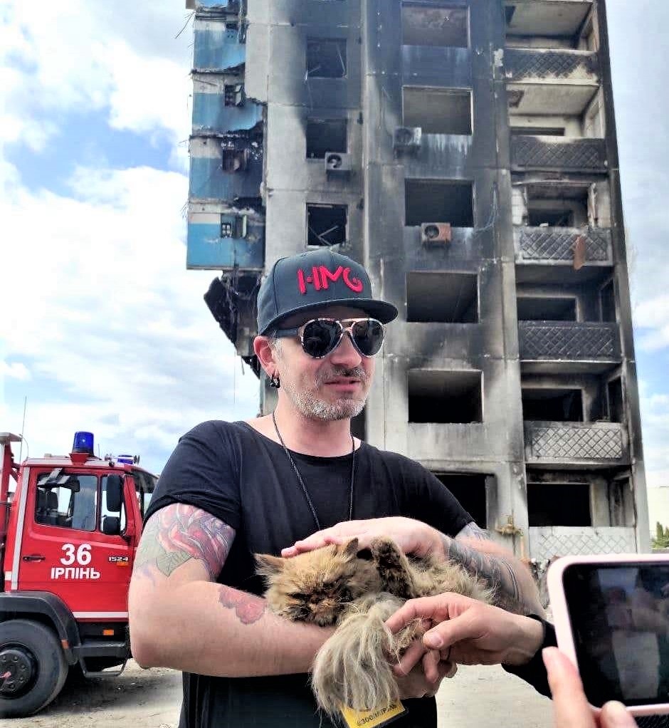 A man in a black t-shirt wearing a ball cap, sunglasses and tattoo on his right bicep and an earring in his right ear holds a tawny, long-haired cat. Behind him is a burnt-out high rise and the front end of a fire truck.