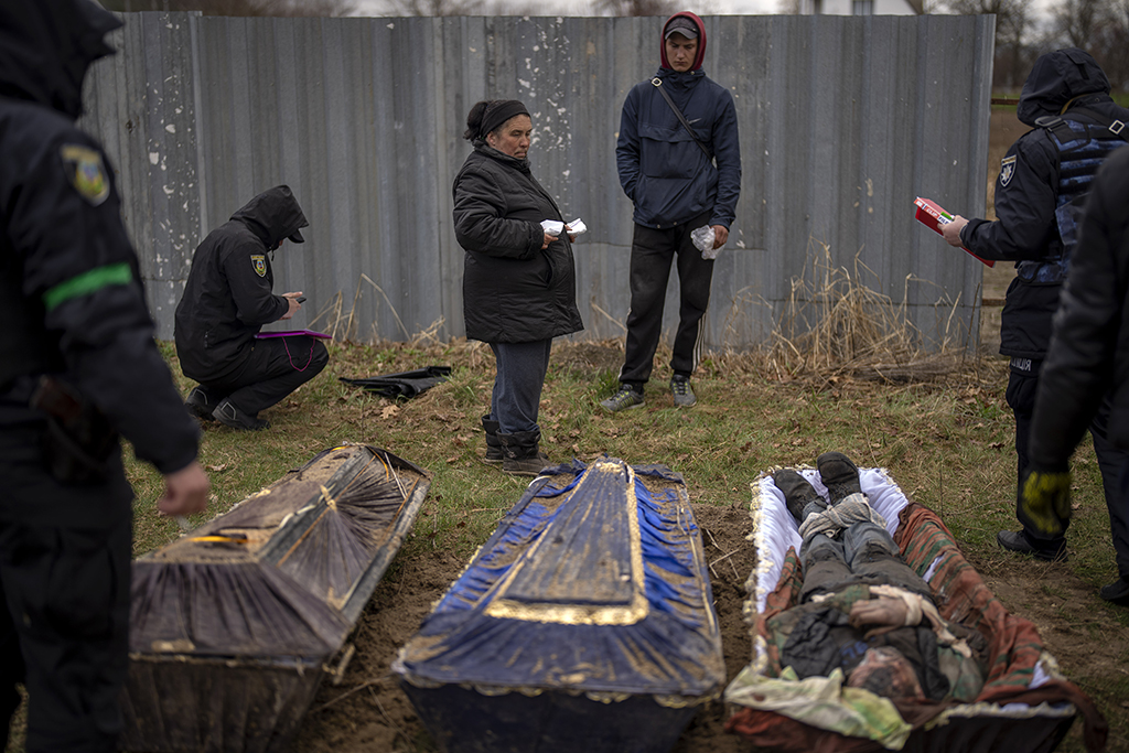 A middle-aged woman stares at a body in an open casket, which is next to three other closed caskets. Around her are people in dark uniforms. 