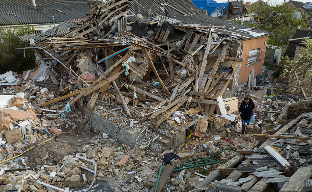 A small, lone figure of woman in a black sweatshirt and jeans walks through the shattered wood and bricks rubble of what used to be a house.