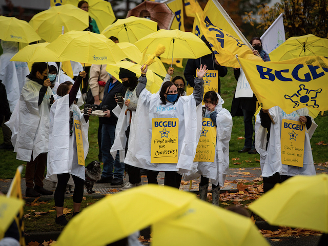 A crowd of workers wearing white rain ponchos and holding yellow umbrellas gathers on a sidewalk and grassy area that is strewn with fall leaves. Signs read things like, “I can’t afford to work for Lifelabs” and “Fair wage for couriers!”  