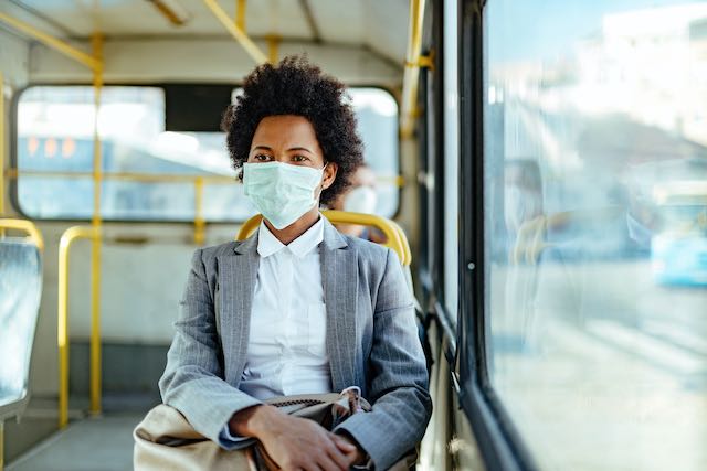 A person with black curly hair is seated on public bus and looking into the distance away from the camera. She is wearing a light green medical mask, a grey blazer, and a white shirt. She is holding a light brown satchel in her lap. 