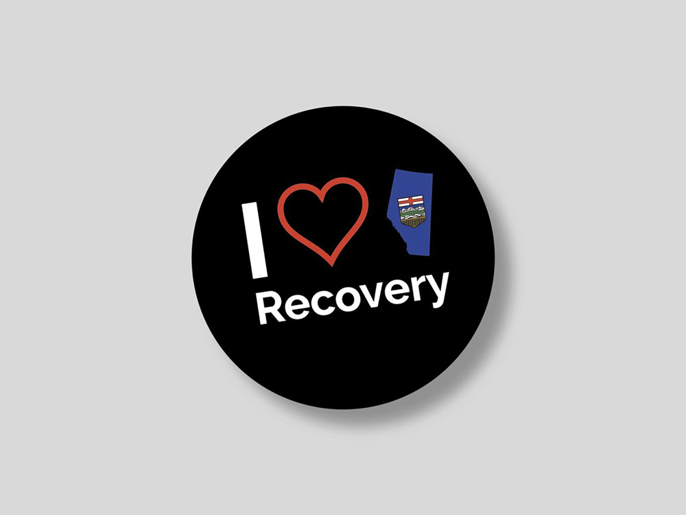 A black circular shape on a grey background reads “I heart recovery” in white text. The heart is an illustrated red outline of a heart next to a blue cut-out of the province of Alberta as it would appear on a map, with its provincial crest at the centre.