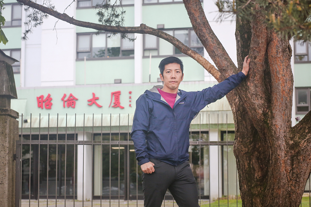 Mike Tan, the vice-president of the Chau Luen Society, leans against a tree. Behind him is his society’s tower for seniors. The tower is painted lime green, and at the entrance are the traditional Chinese characters for Chau Luen Society in red.
