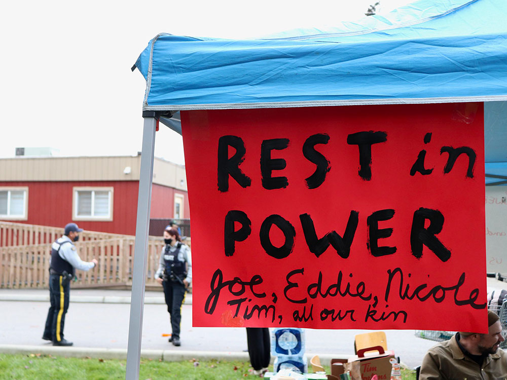A handmade red sign hangs from the roof of a folding outdoor tent with a blue tarp. Hand-painted letters read, “Rest in Power Joe, Eddie, Nicole, Tim, all our kin.” A person in a toque and tan jacket is sitting to the right of the frame under the tent. Two police officers in medical masks are speaking with each other on the street in the background.