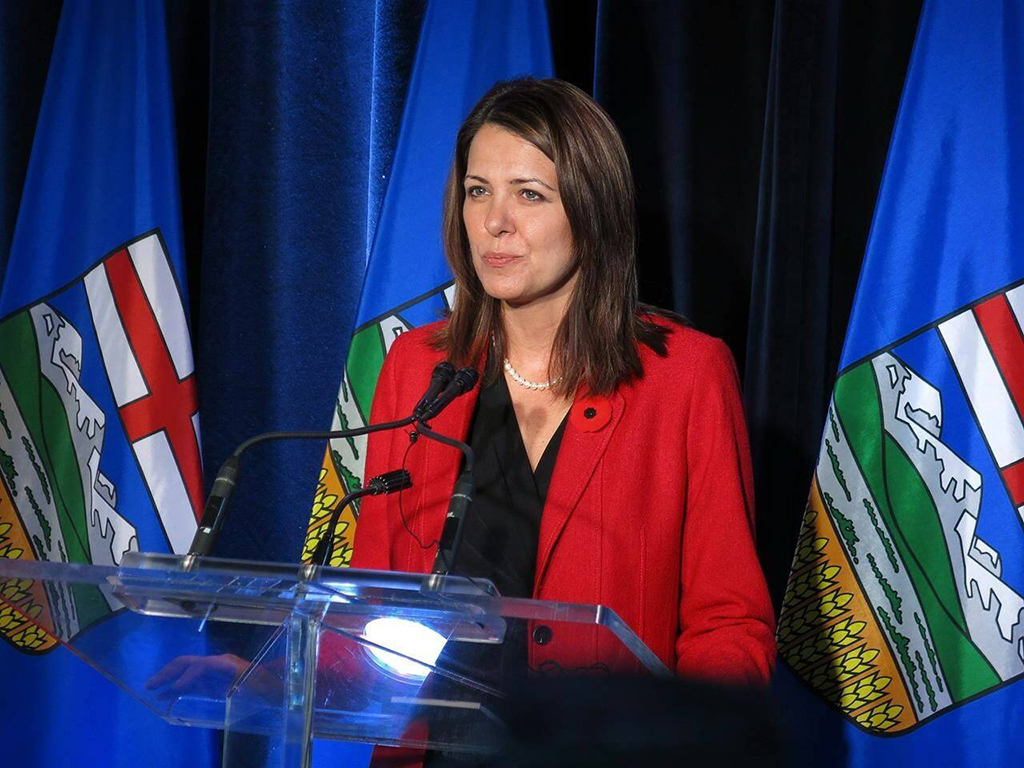 Danielle Smith stands before a podium, flanked by two flags, in a red blazer.