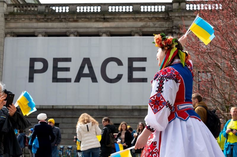 A person in traditional Ukrainian clothing holds handfuls of small blue and white Ukrainian flags at a pro-Ukraine rally in downtown Vancouver on March 27, 2022. The rally took place at the Vancouver Art Gallery; the gallery’s exterior wall holds a large sign that says ‘Peace’ to commemorate its exhibit at the time showcasing the work of Yoko Ono. People are rallying for Ukraine in front of that sign.