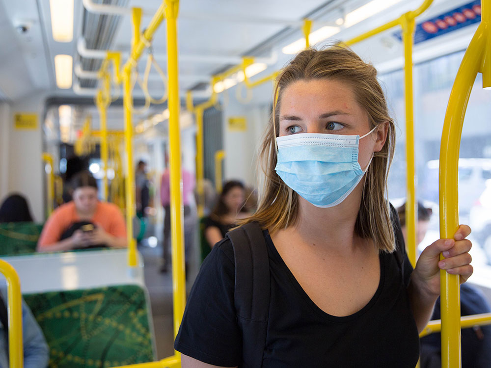 A young woman stands holding a yellow pole on public transit. She has blonde hair and is wearing a blue medical mask.