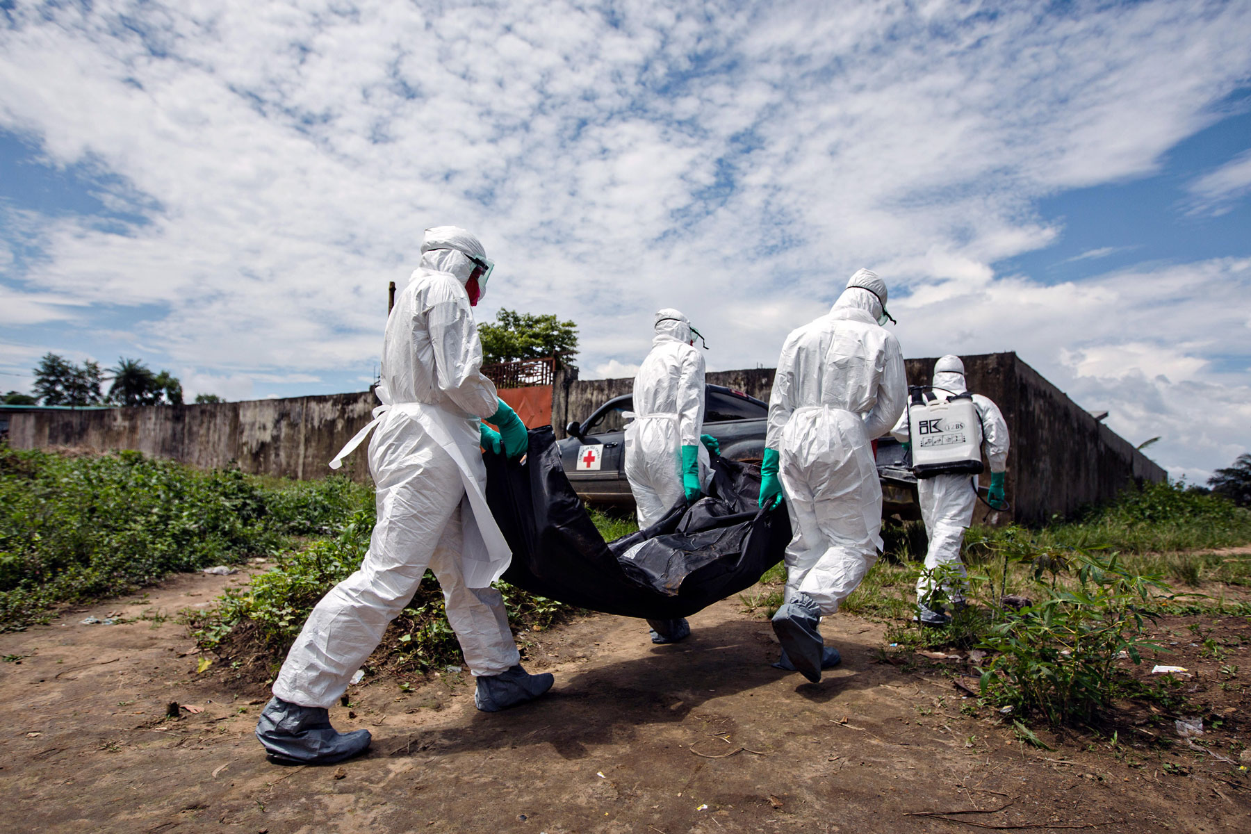 Four people walking in white medical gear, three of which are carrying a wrapped body.