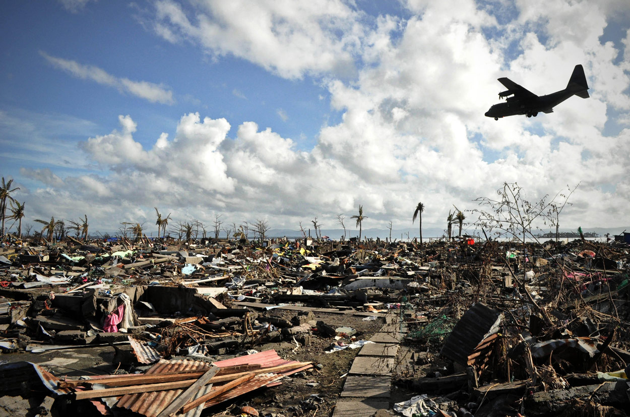 A large military plane flies overhead piles of rubble and blown-over trees.