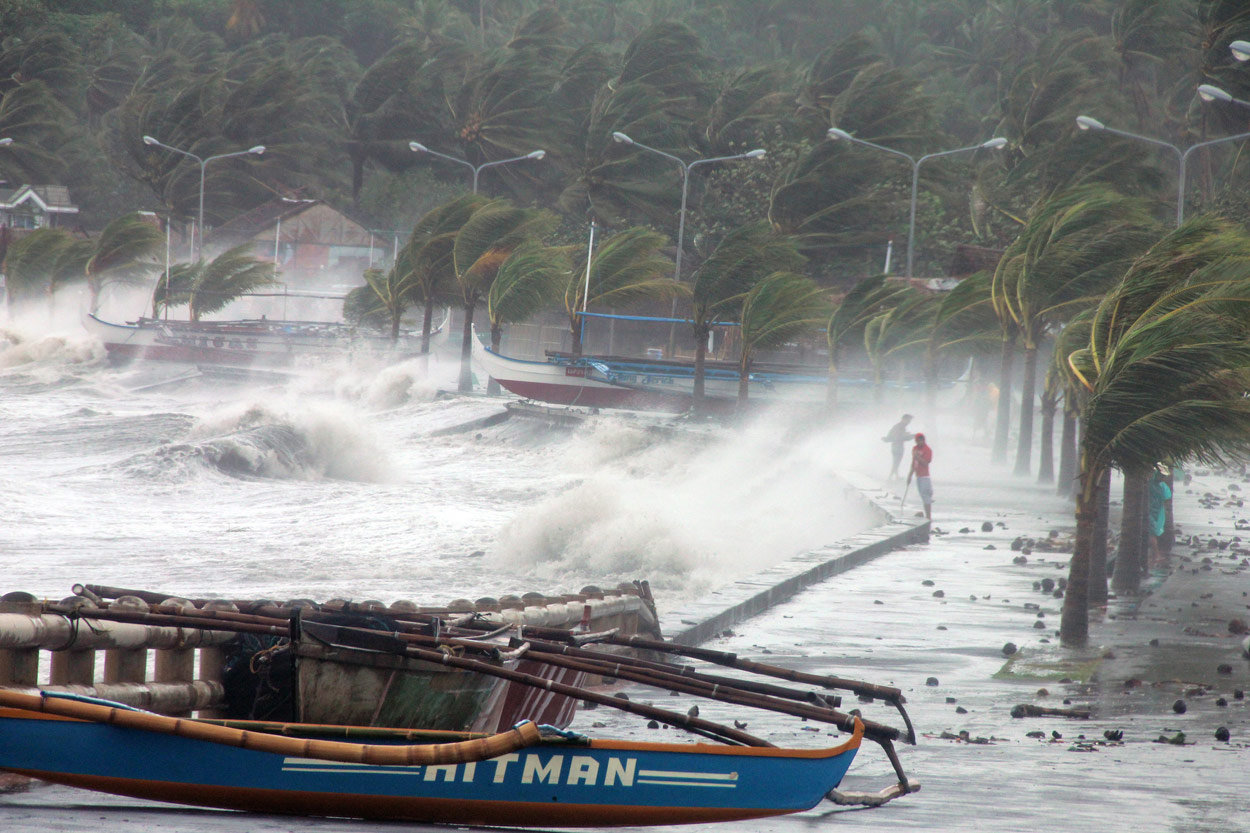 A boat in the foreground, very strong winds blowing palm trees, and waves crashing ashore with residents standing nearby.
