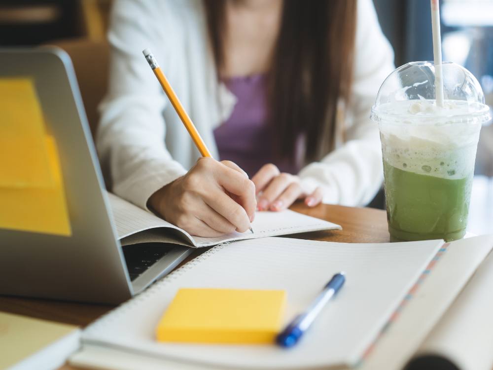 A student writes with a pencil in a notebook. We can’t see their face, but they are wearing a white cardigan over a purple stop. A silver laptop with yellow sticky notes on it, another notebook, a yellow sticky notepad and a frothy green beverage crowd their desk. 