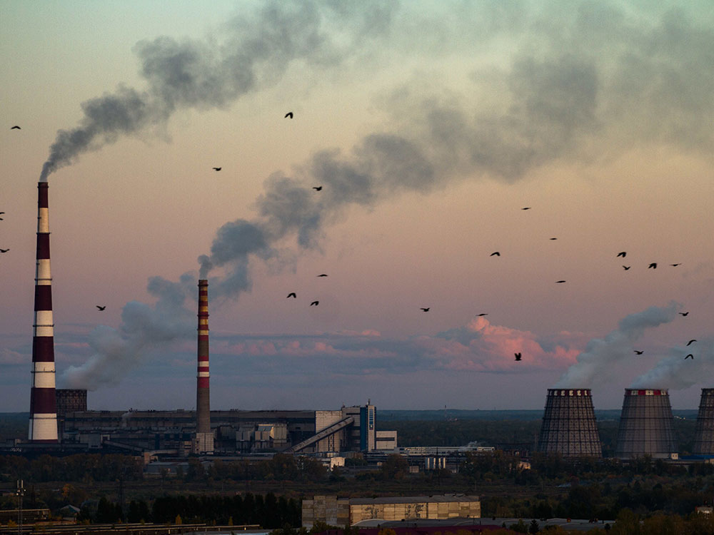 It’s twilight as birds fly by industrial smokestacks in Omsk, Russia. The tallest smokestack, striped in black and white, is to the left of the frame. To the right is a trio of shorter brown smokestacks.
