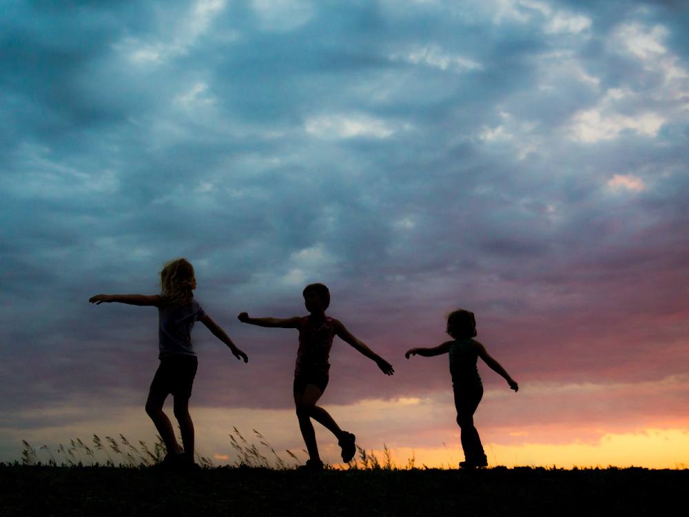 Three children walk with arms outstretched. Behind them is a wide, expansive sky with clouds and a sunset fading from red and pink into blue. 