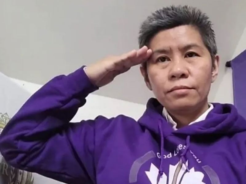 An unsmiling woman with a short haircut wearing a purple hoodie salutes while facing the camera.