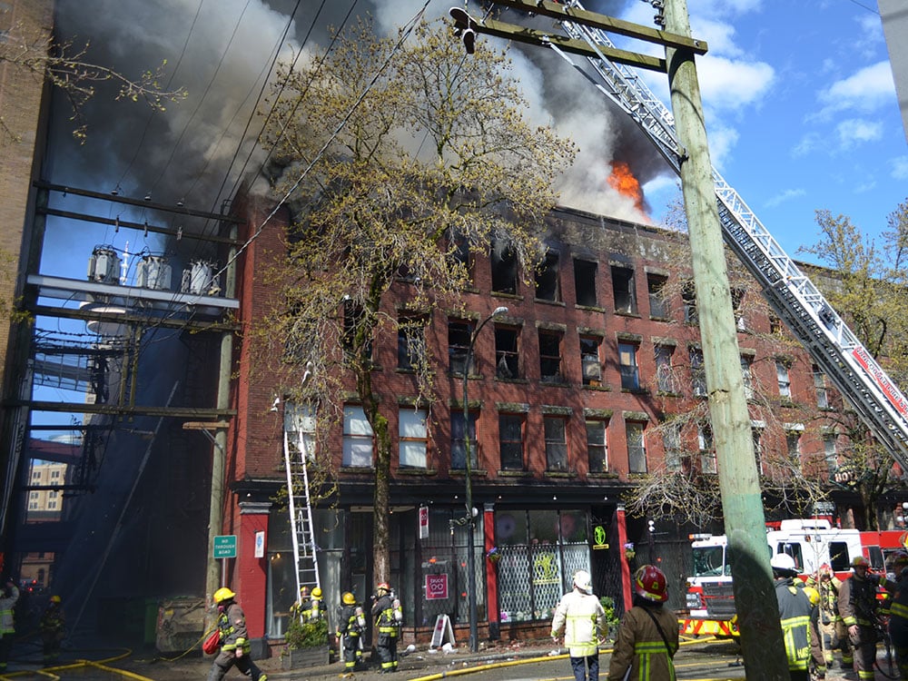 Firefighters, some with a ladder, gaze at a four-storey brick building. Some windows are blackened and broken, and flames and smoke surge from the roof, all under blue skies.