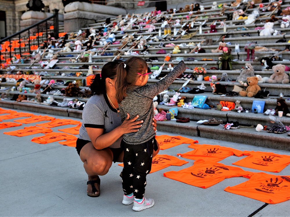A woman crouches and gently hugs her child, who is pointing towards orange shirts, shoes, flowers and messages on display on a set of stairs outside the legislature.
