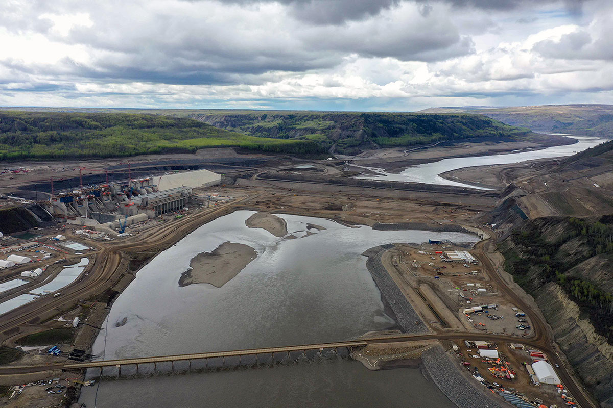 An aerial view shows the Peace River and its valley. A new bridge crosses the river in the foreground. Above it, there is an earth dam and on the left a concrete structure that will house the power-generating equipment. Above the dam, the river valley is flanked with wide swaths of dirt where trees have been logged in advance of flooding.