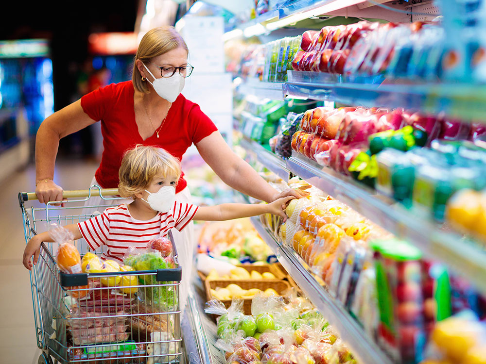 A parent shops in a grocery store with her young child riding in a shopping cart. They are both reaching into a produce fridge to the right of the frame. They are both wearing white medical masks.