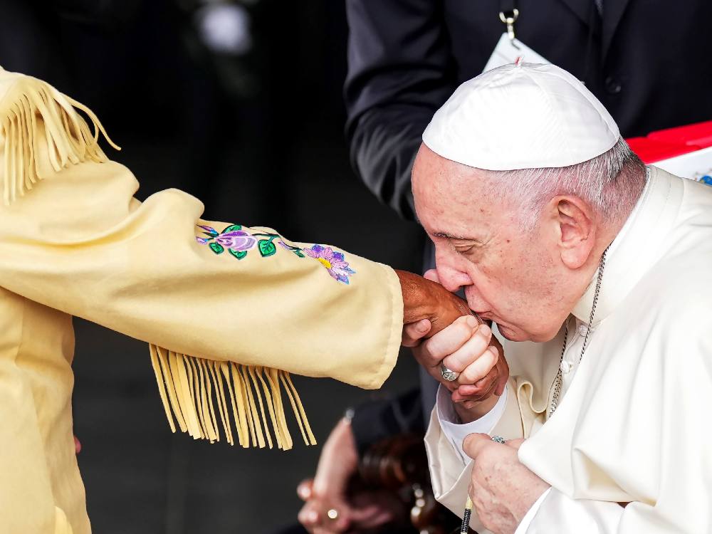 An outstretched arm wears embroidered buckskin. Pope Francis kisses the wearer’s hand.