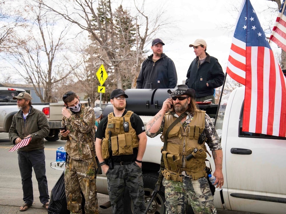 Six men stand in front of and in the bed of a silver pick-up truck decorated with American flags. Three men in front of the truck are clad in camouflage patterned clothing reminiscent of the military. Some are holding weapons and one is using a cell phone.