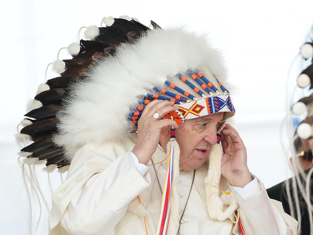 An older white man in white robes is adjusting a headdress with a beaded band across his forehead and long feathers rising above his head.