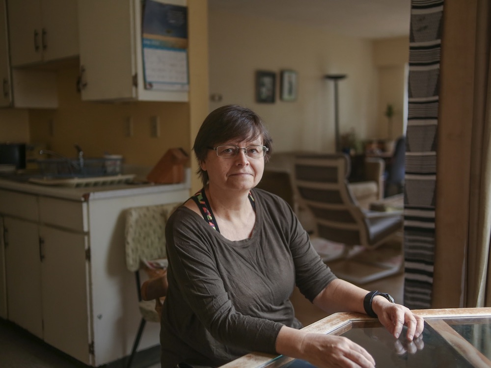 A woman in her mid-50s sits at the kitchen table of her roomy apartment. Behind her on the left is the kitchen and behind her on the right is her living room. It looks like a cozy, lived-in space.