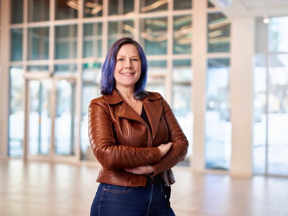 A smiling woman with dark hair with purple highlights stands with arms crossed. She is wearing a short brown leather jacket and blue jeans.