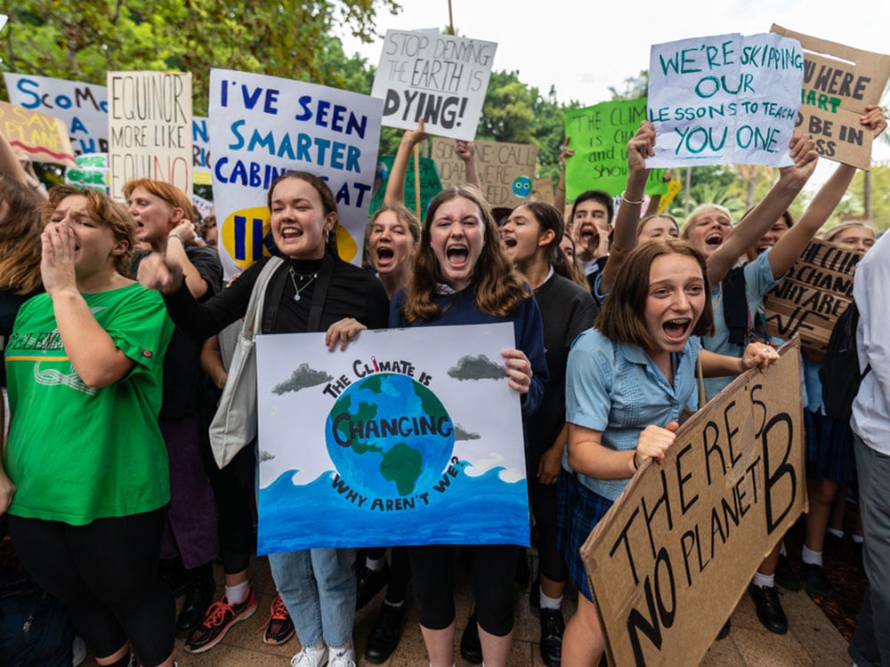 A large group of student protestors gather with placards reading several different things, including: “There’s No Planet B,” “Stop Denying the Earth is Dying,” and “The Climate is Changing — Why Aren’t We?”
