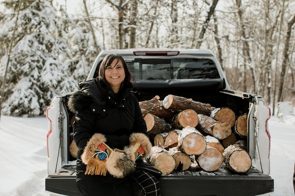 A woman sits on the open gate of a pick-up truck, smiling. Behind her, there is a cord of firewood in the bed of the pickup truck.