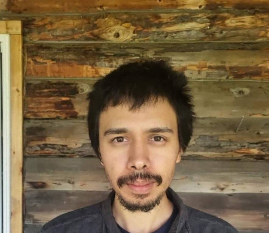 A headshot of a man with a short beard and mustache. He’s standing in front of a log cabin.