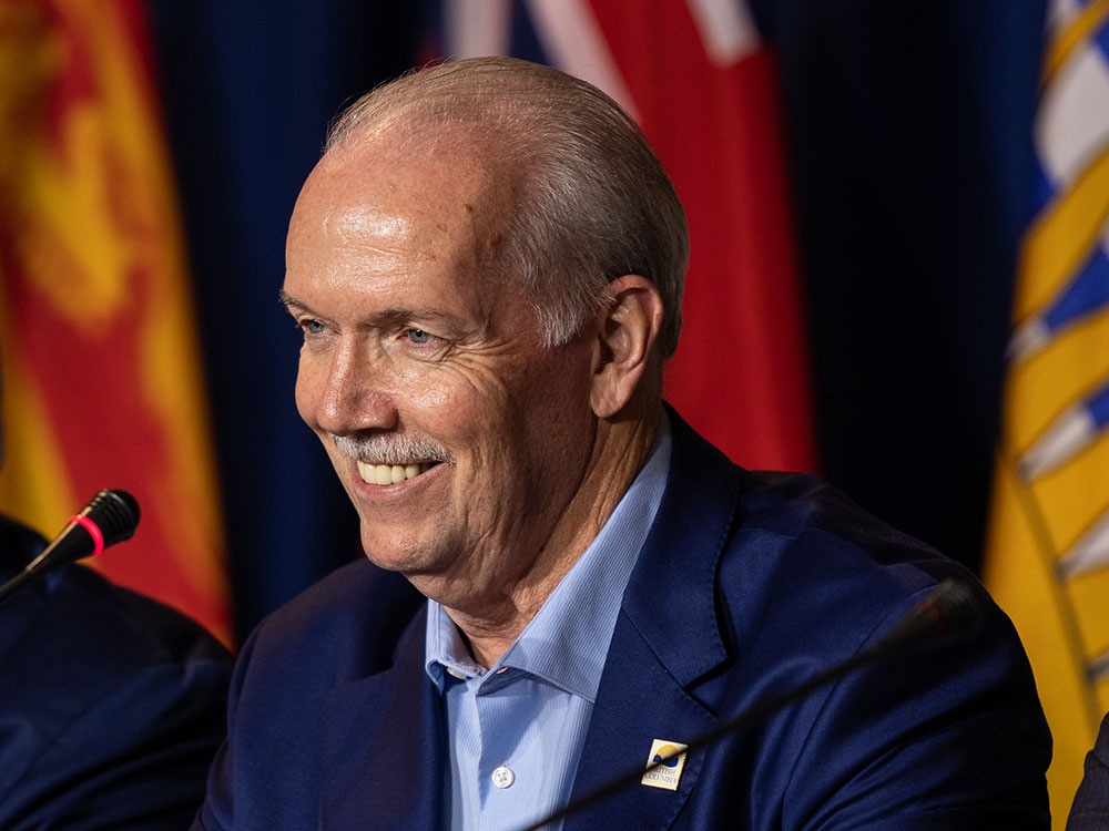 The smiling face of B.C. Premier John Horgan, a late middle aged white man, balding with grey hair combed back, wearing a blue suit, is shown leaning forward towards a microphone with a B.C. flag out of focus behind him.