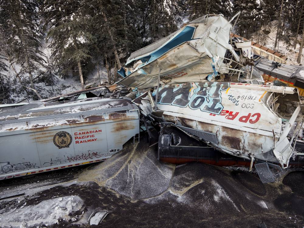 Train cars are piled up in a mess of twisted metal and spilled contents. It’s winter, and the ground and surrounding trees are covered in a light blanket of snow.