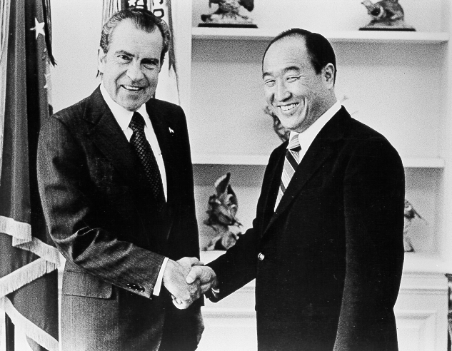 Two men in suits shake hands with US flag behind.