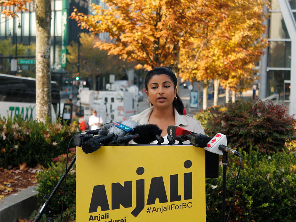 With trees in fall colours in the background, a woman of colour in an off-white jacket stands behind a podium with media microphones.