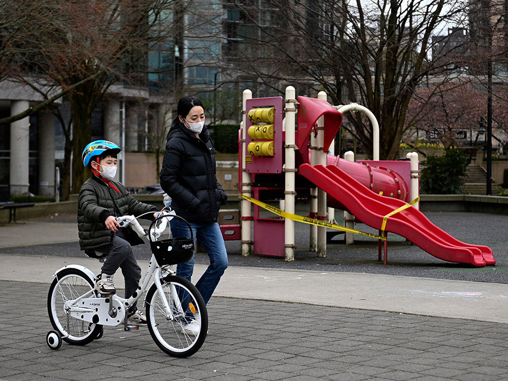 An Asian woman is walking beside a young boy riding a bicycle with training wheels in an urban environment. They are both wearing face masks. It is a grey day. Towers and cherry blossoms are visible in the background. The pair is passing a small empty playground with yellow tape on its red slide and climbing apparatuses. 