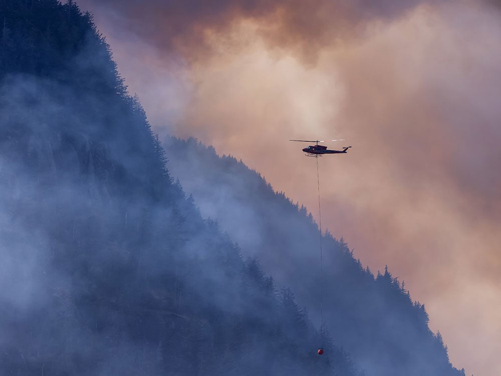 A BC Wildfire Service helicopter flies over a forest fire and smoke on the mountain near Hope during a hot sunny summer day.