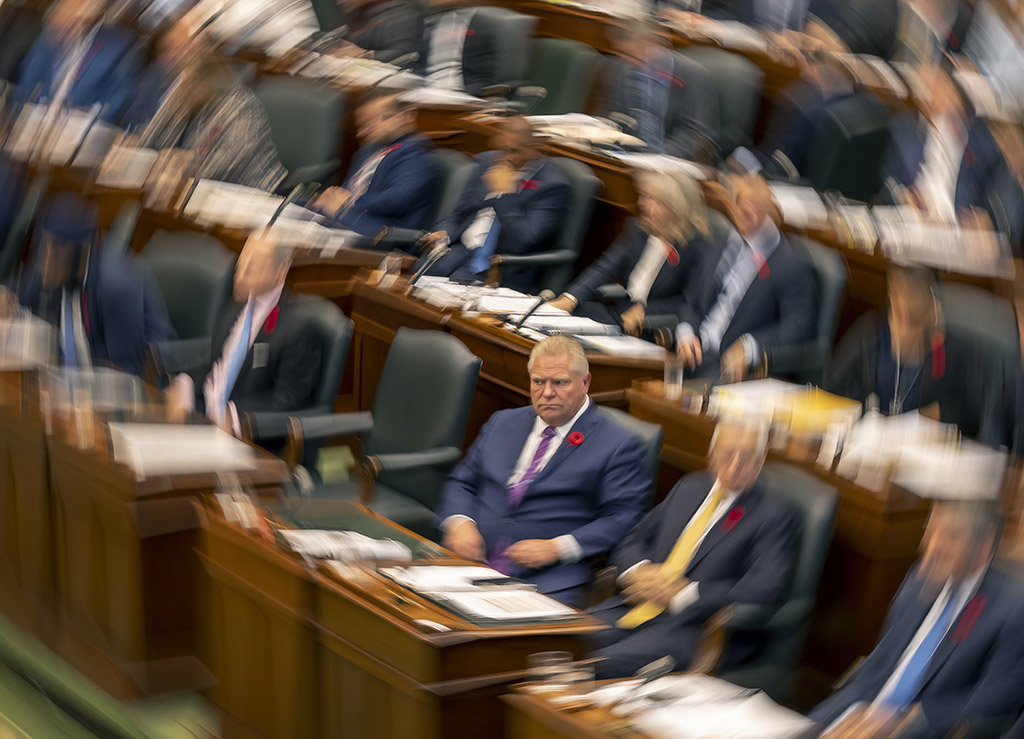 A stylized photo shows the government benches in the Ontario legislature, almost entirely men in suits and ties. The photographer has created a swirling image, where only Premier Doug Ford — a burly, light-haired man in a white shirt, red tie and a blue suit with a poppy — is the only figure in focus.
