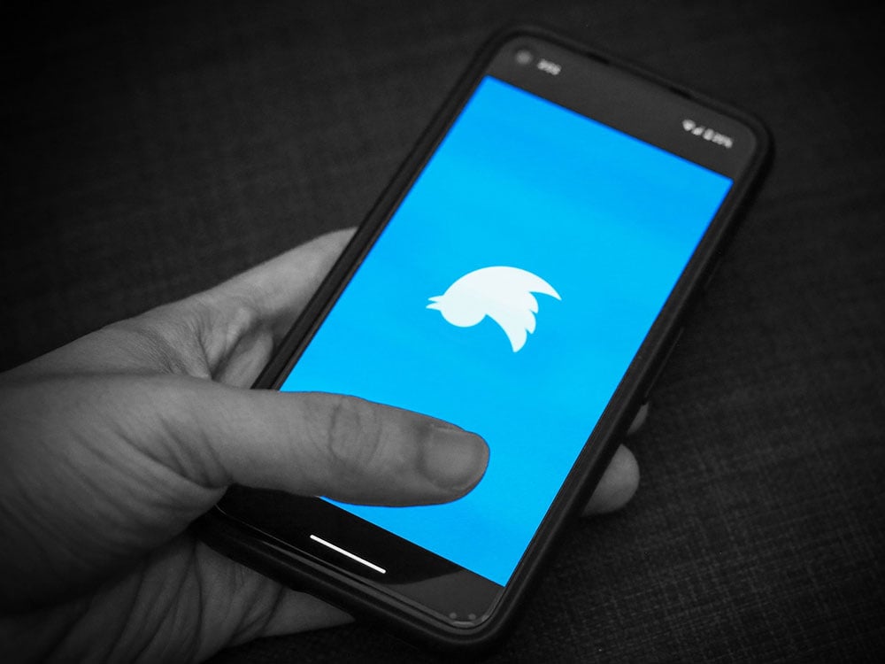 A closeup of a hand, in black and white, holding a phone. It’s screen shows a blue field and an upside down Twitter logo. 