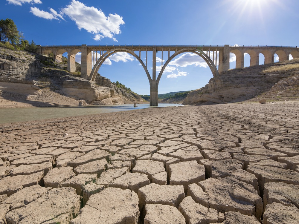 Under a brilliant blue sky, a tall bridge stretches gracefully between two sides of a canyon. Beneath it the riverbed is dry and cracked, with a small amount of water.