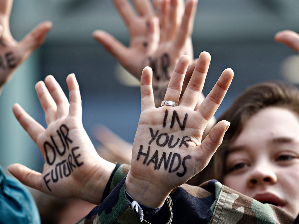 High school and university students stage a protest against the climate policies of the Belgian government. The camera is zoomed in on a youth who has written “Our Future Is In Your Hands” on their palms, and is facing their palms to the camera. 