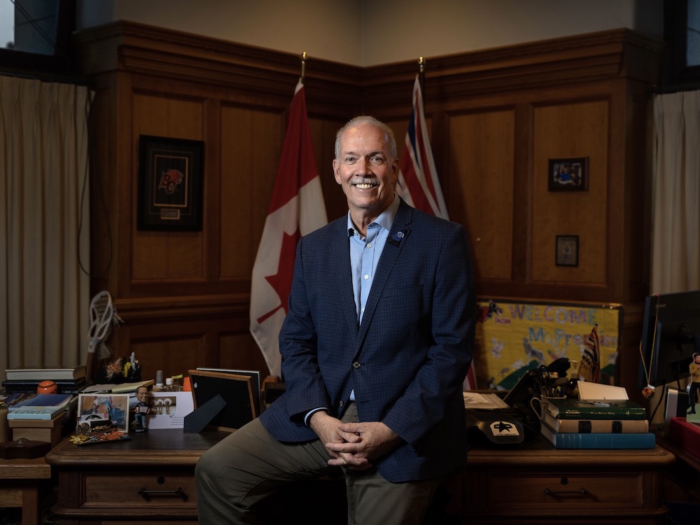 Premier Horgan partially stands, partially sits on his desk in the legislature. He is smiling at the camera. Behind him, in the corner of his office, are BC’s flag and the Canadian flag.