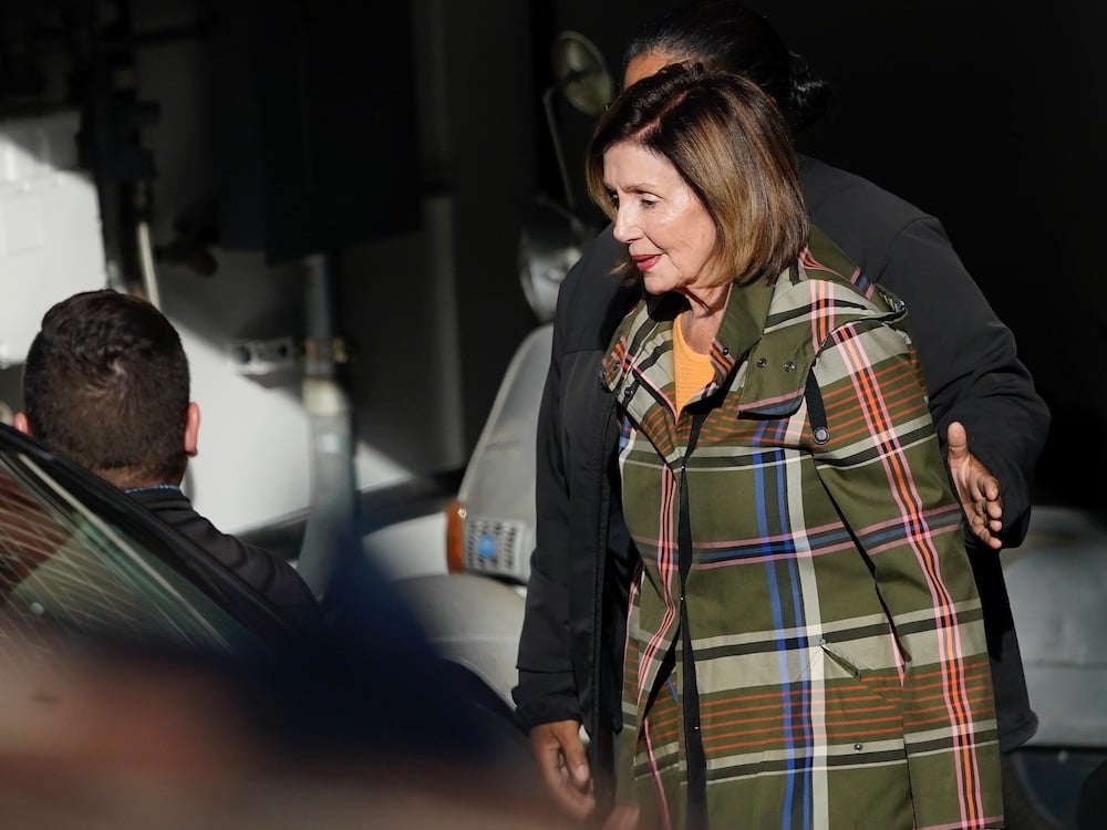Nancy Pelosi is walking towards the left of the frame. The side of her face is visible; she has short brown hair and is looking down to the vehicle front of her. She is wearing a green jacket with a wide plaid pattern over a yellow shirt. Behind her, a security guard in a black jacket is escorting her down the street. We can’t see their faces. The background is dark with shadows.