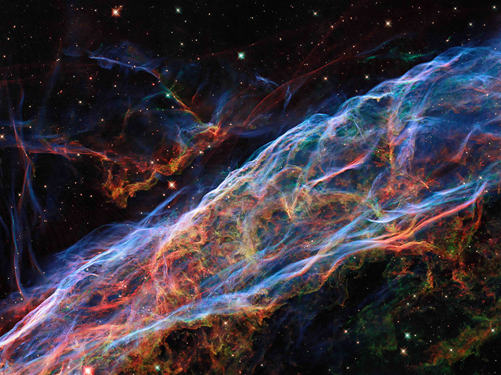 This image of the Veil Nebula depicts an oblong translucent shape bisecting the frame from the bottom left corner to the top right. According to NASA/ESA, the image picks up enhanced details of emissions from doubly ionized oxygen (seen here in blues), ionized hydrogen and ionized nitrogen (seen here in reds). The nebula is set against a black starry sky.