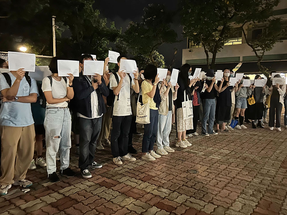 Young adults dressed in casual streetwear (T-shirts, denim, sneakers) stand next to each other in rows, holding blank pieces of white paper in front of their faces to commemorate a recent deadly fire at the Chinese University of Hong Kong in Hong Kong. They are standing on an outdoor plaza with a concrete tiled walkway. The sky is dark and most people’s faces are either covered by papers or medical masks. 