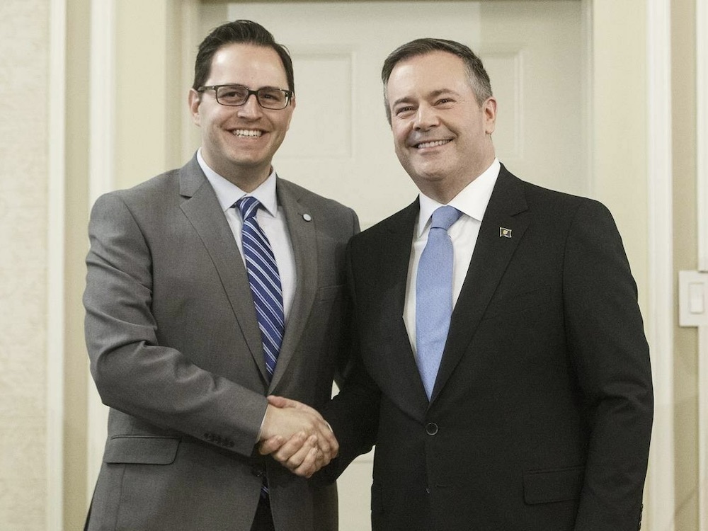 Two white middle aged men with dark hair wearing suits and ties shake hands.