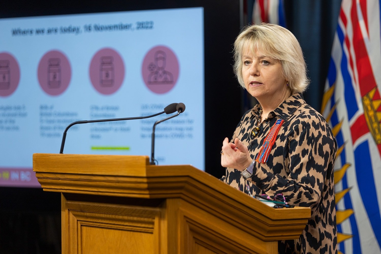 A woman with short blonde hair and a leopard-print button-down shirt stands at a wooden podium behind a pair of microphones. She is standing to the right of the frame in front of red, white and blue British Columbia flags. Behind her is a PowerPoint slide containing an infographic with light red and white colours conveying public health information.