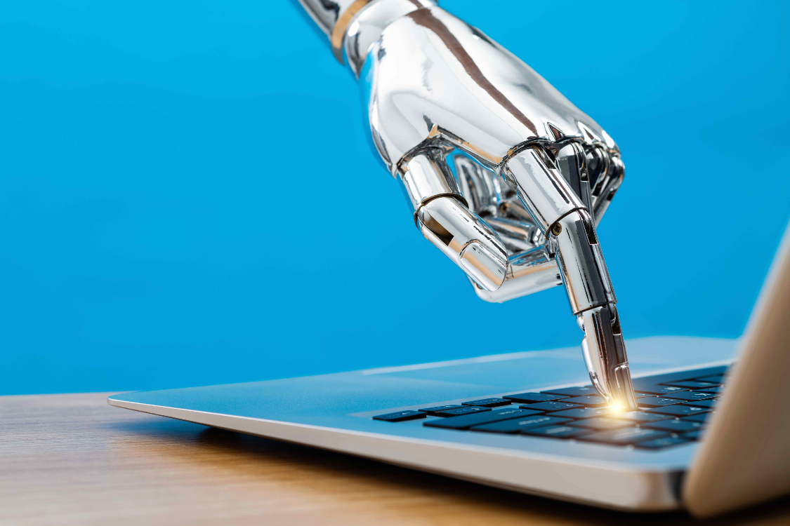 A silver robot hand places its index finger on a the black keyboard of a grey MacBook Air laptop on a wooden table. The tip of the finger glows gold. The background is blue.