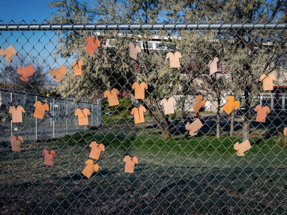 Numerous paper orange shirts are arranged on a chain link fence in front of a public school in Kamloops, B.C. in October 2021. The school is visible in the background behind a stand of trees and a green field. It’s a sunny autumn day and the sky is blue.