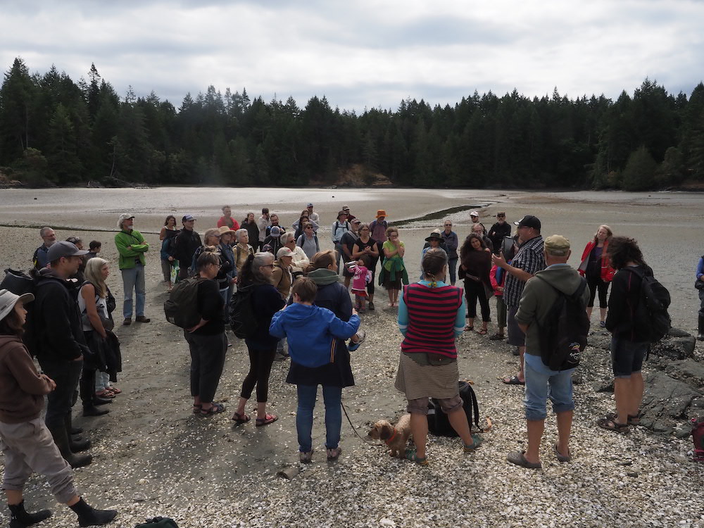 A group of about forty people stands in a circle on a beach.